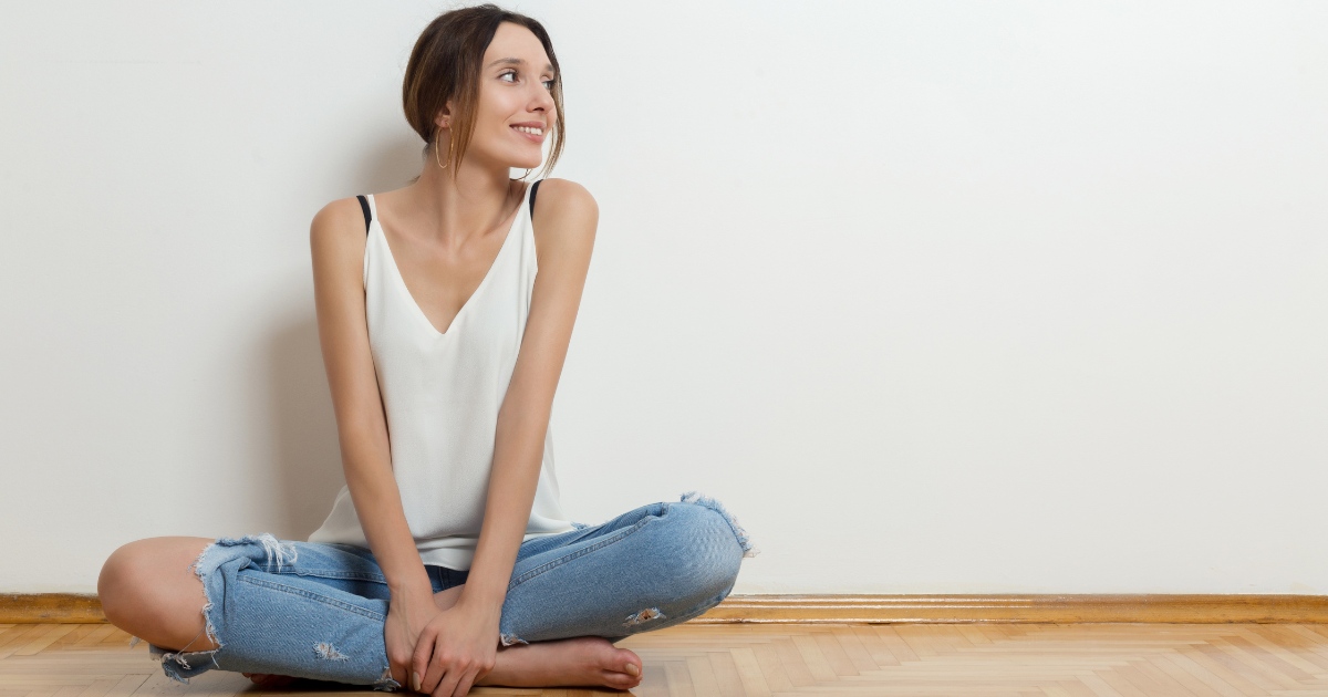 A woman sitting cross-legged on a wooden floor against a plain white wall. She is wearing a white sleeveless top and ripped blue jeans, smiling and looking to her right with a relaxed, cheerful expression.