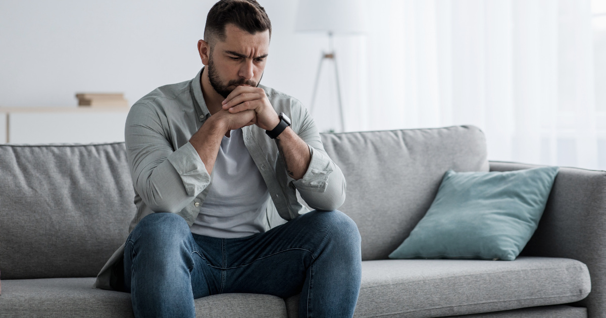 A bearded man sits on a gray couch, leaning forward with his hands clasped near his face, looking thoughtful and concerned in a bright living room.
