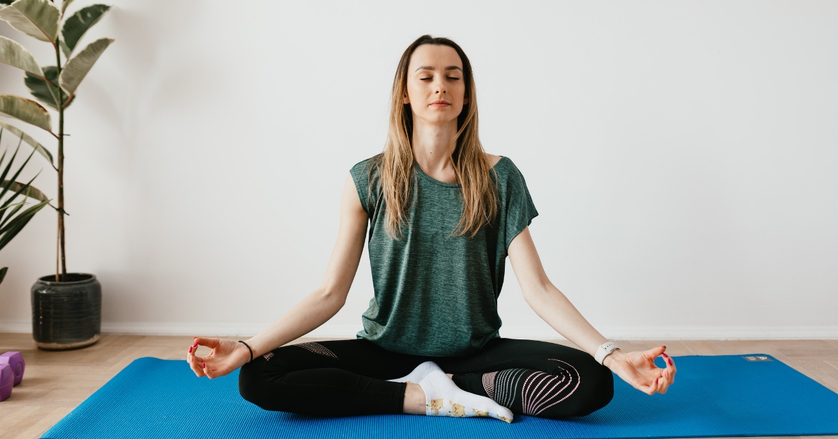 A woman sits cross-legged on a blue yoga mat with her eyes closed and hands resting on her knees, practicing meditation in a calm, minimalist room.