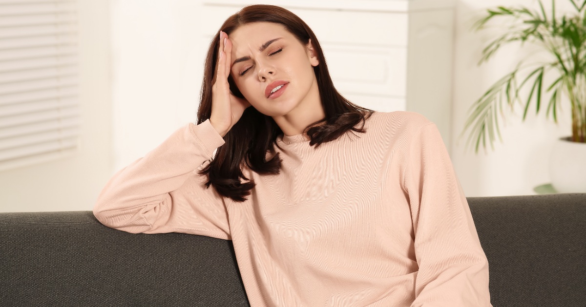 A woman sits on a couch with her eyes closed, pressing her hand to her temple as if experiencing a headache, in a bright living room with a plant in the background.